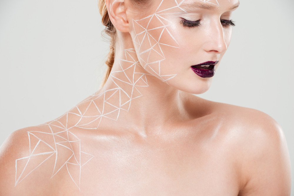Cropped portrait of beauty woman with body art A woman with dark lipstick with geometric shapes drawn on her from her shoulder, up her collarbone, onto her forehead.