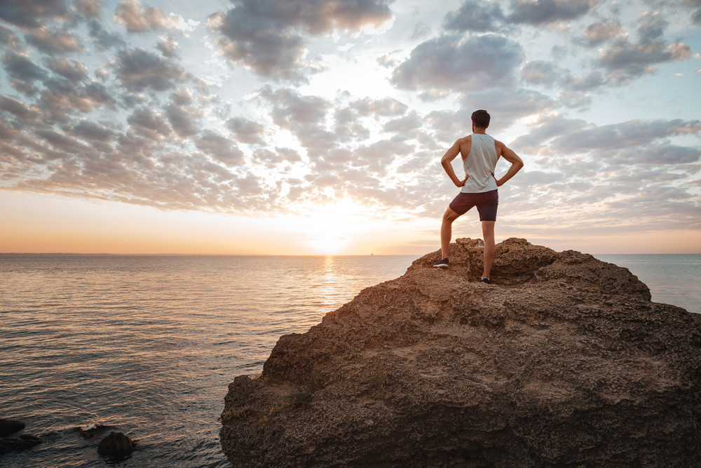 Young sportsman standing on the mountain rock by the sea A man in a tank top and shorts standing on a large rock overlooking an ocean sunset.